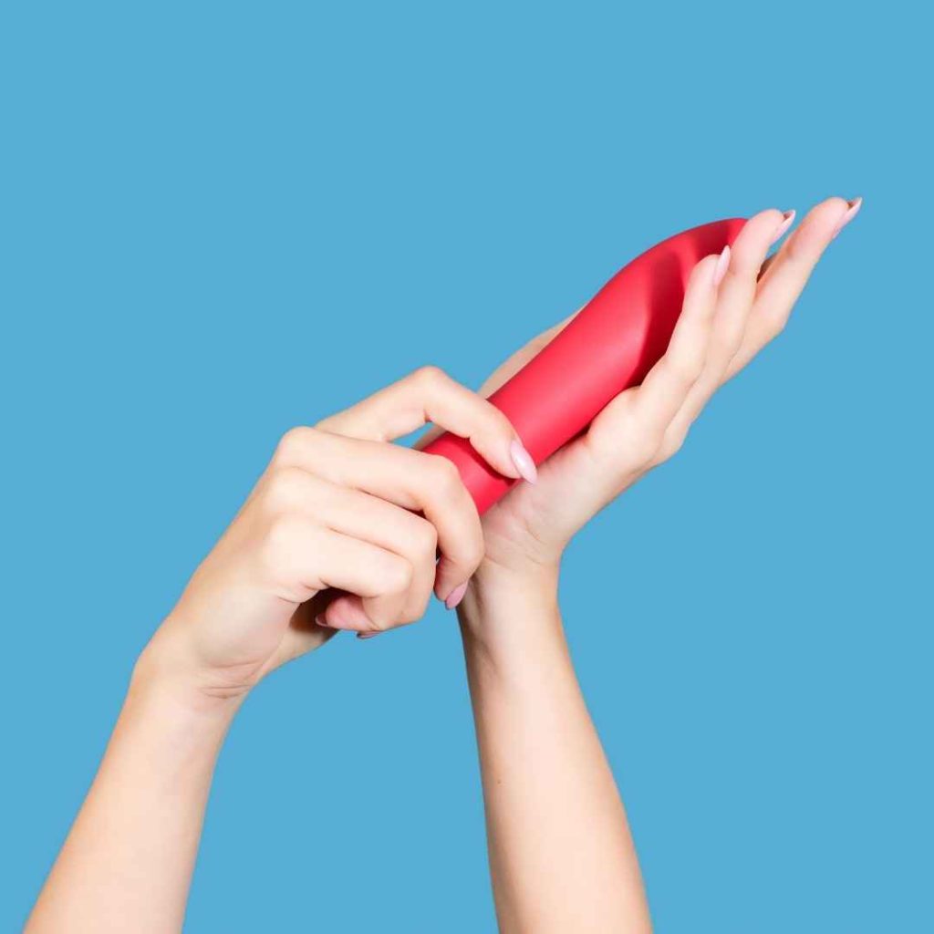 Hånd holder rød vibrator mod blå baggrund, moderne silikone sexlegetøj til klitorisstimulering og solo leg