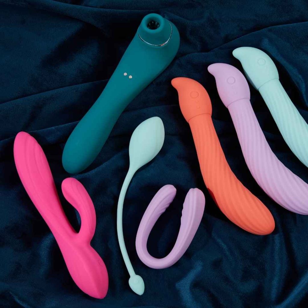 Forskellige farverige vibratorer og par vibratorer placeret på mørkt stof, sexlegetøj til klitorisstimulering og vaginal nydelse