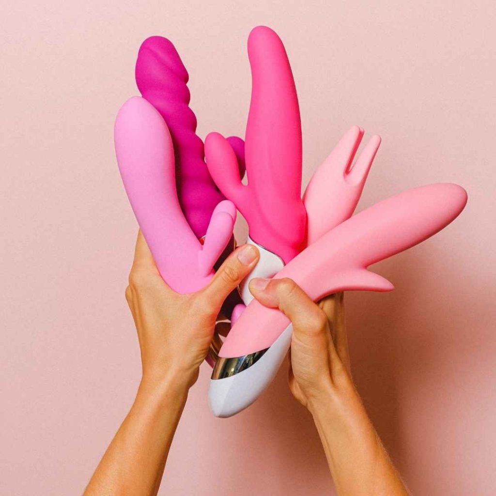 Hænder holder flere pink og lilla vibratorer samlet, moderne sexlegetøj til klitorisstimulering og vaginal nydelse