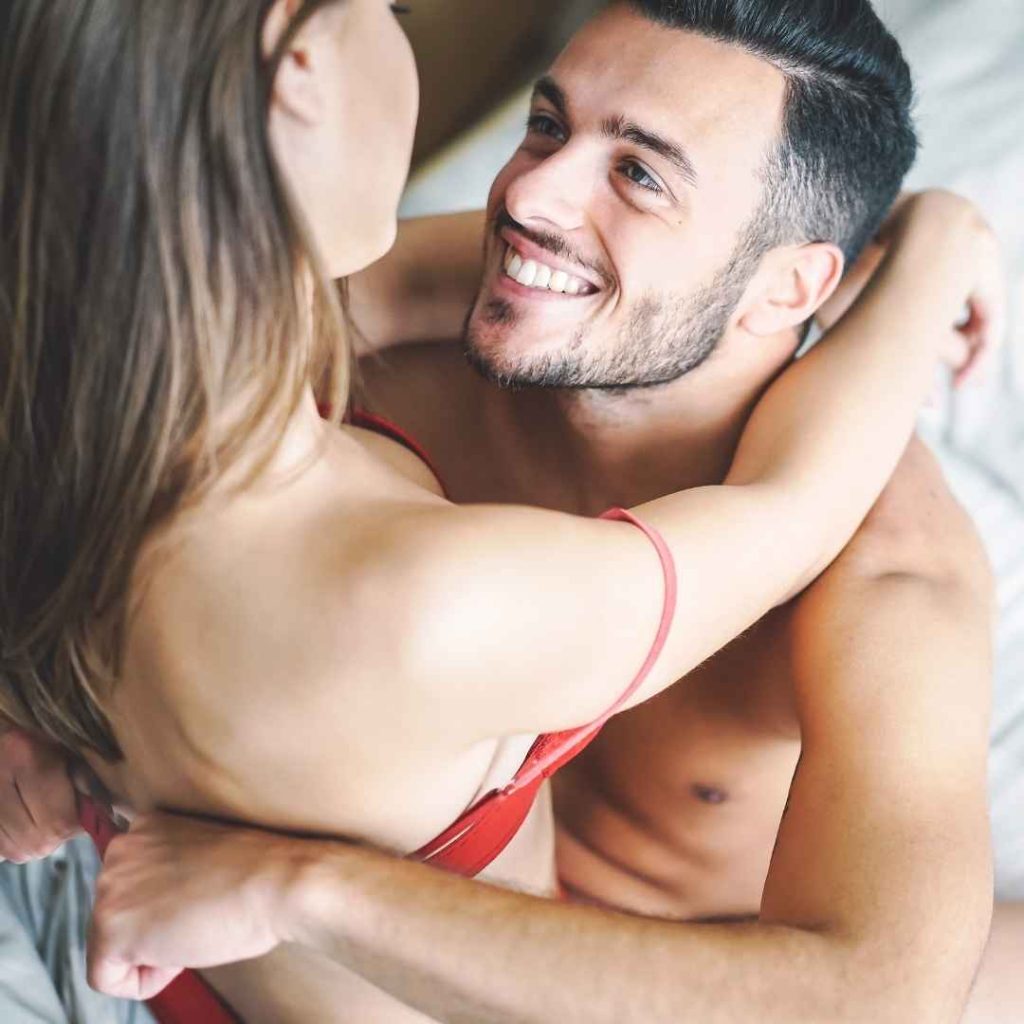 Smilende par omfavner hinanden i soveværelset, intim stemning og fokus på kærlighed og seksuel nærhed