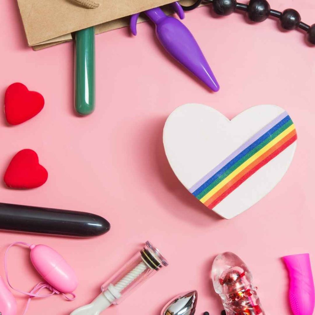 Forskellige vibratorer og dildoer arrangeret på lyserød baggrund med regnbuehjerte, sexlegetøj til solo leg og par i forskellige former og farver