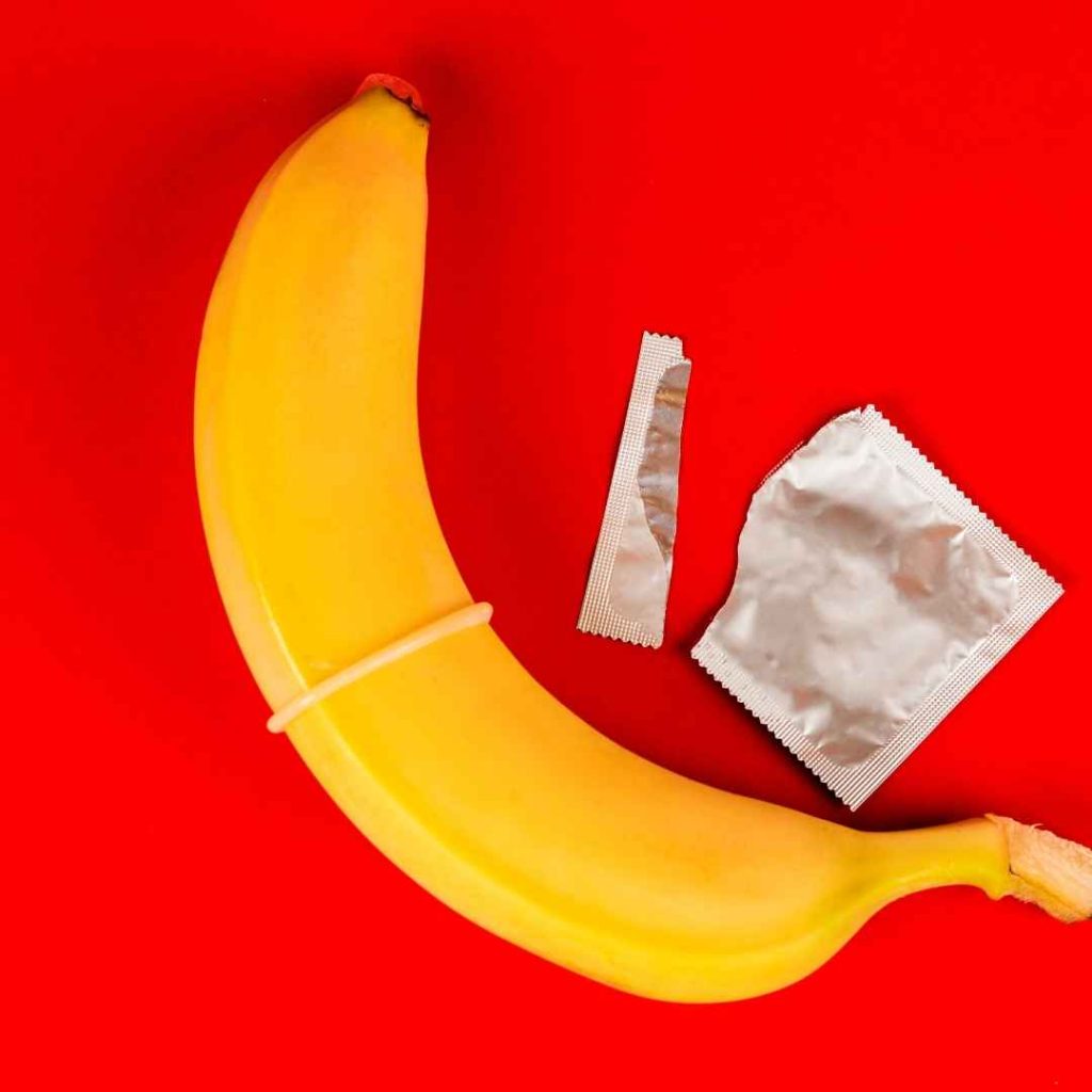 Gul bananformet vibrator på rød baggrund ved siden af kondom og pakke, sjovt sexlegetøj med frugt design til intim stimulering